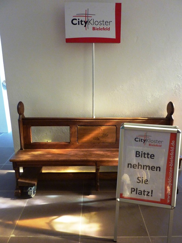 Auf dem Foto sieht man eine Kirchenbank und ein Schild, auf dem "Bitte nehmen Sie Platz" steht.

Foto: CityKloster Bielefeld